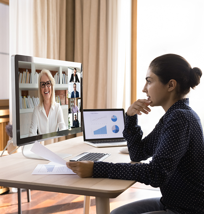 Barrister Suites Office Lease A woman sits at a desk with papers, looking at a computer screen where she is on a video call with five people. A laptop nearby displays charts and graphs. The room is bright with natural light.