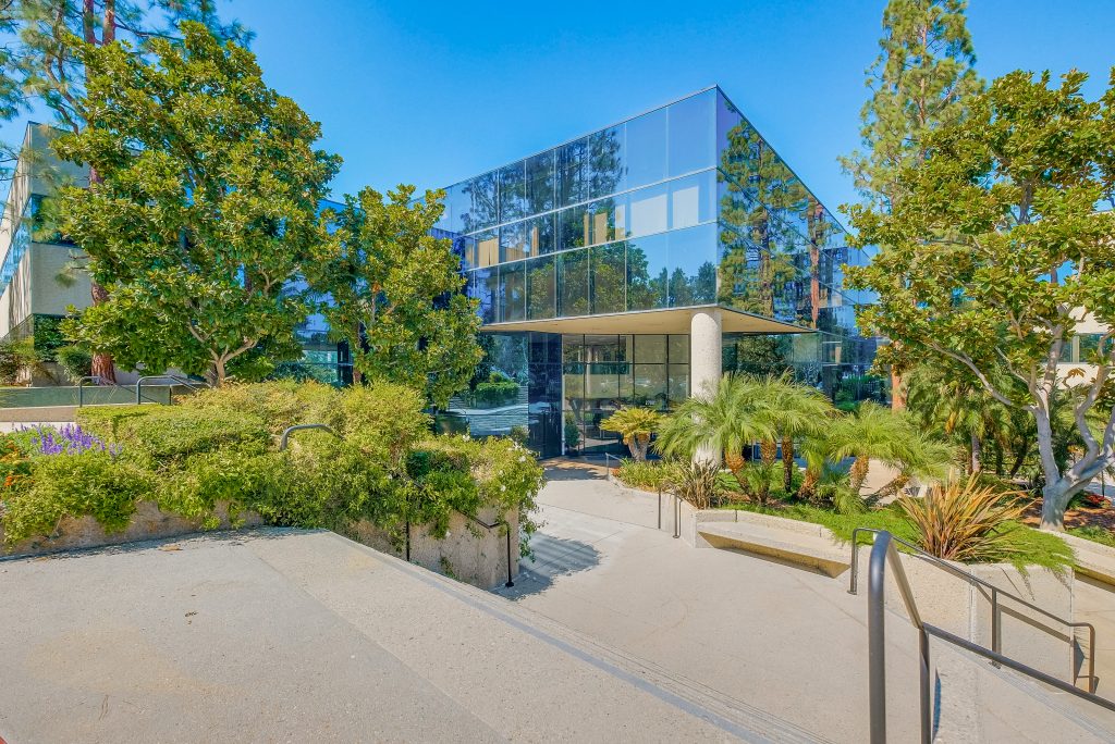 Barrister Suites Office Lease A modern glass office building surrounded by lush green trees and landscaping, with a pathway and steps leading to the main entrance under clear blue skies.