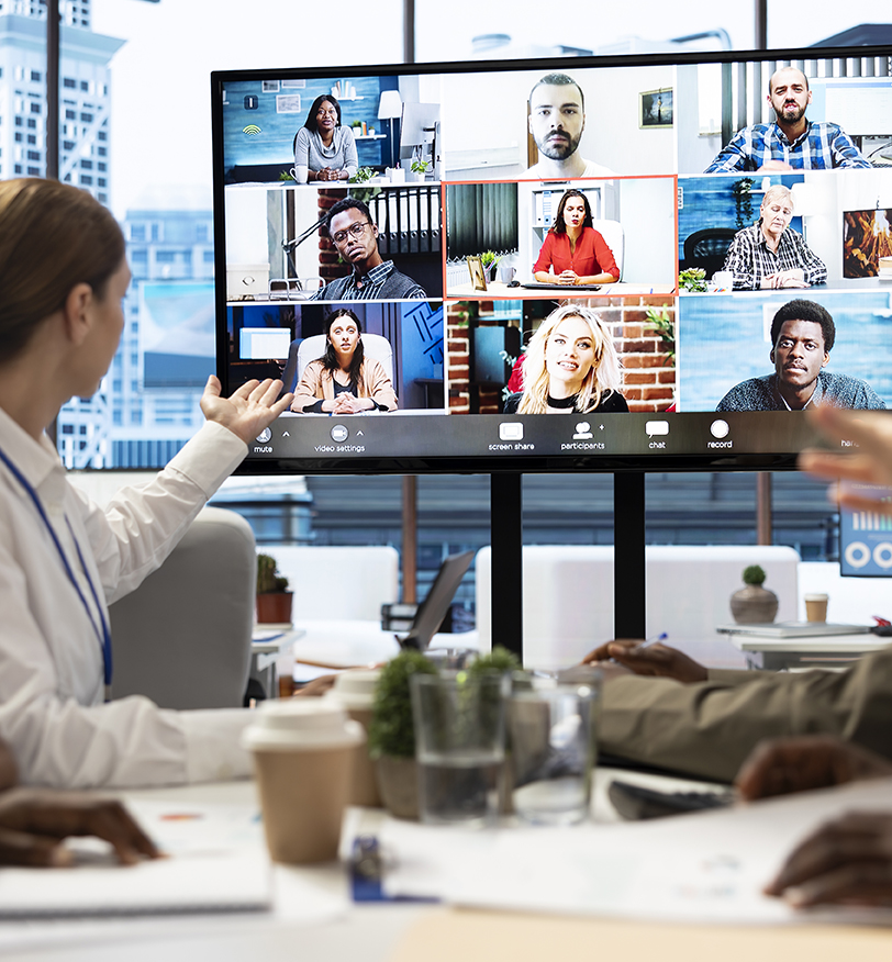 Barrister Suites Office Lease A group of people in an office sit at a table, participating in a video conference call with multiple participants shown on a large screen in front of them. Laptops, coffee cups, and papers are on the table.