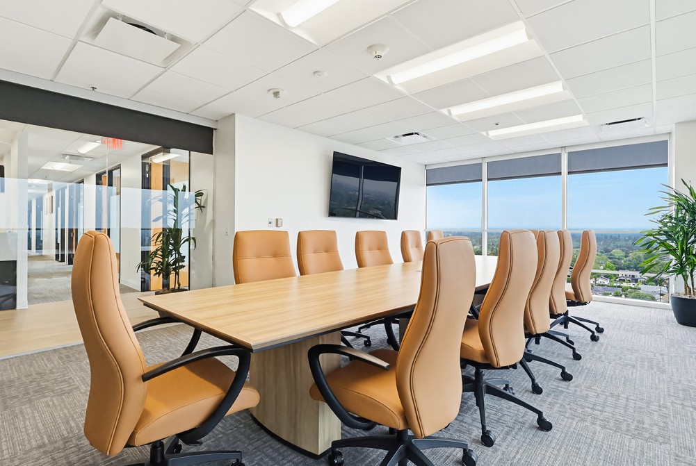 Barrister Suites Office Lease Modern conference room with a large wooden table surrounded by tan office chairs, large windows with city view, a wall-mounted TV, and several green plants. Bright and spacious with natural light.