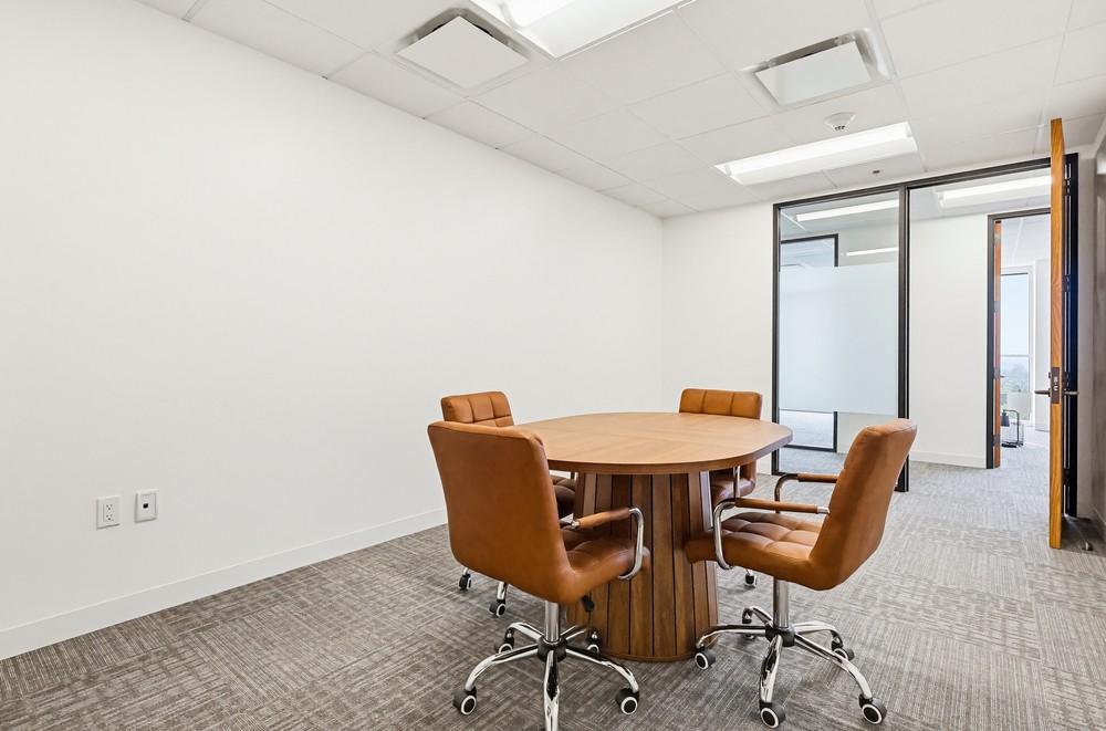 Barrister Suites Office Lease A small modern conference room with a round wooden table and four brown leather chairs on wheels, beige carpet, white walls, and a ceiling with recessed lighting. A glass door leads to another office area.