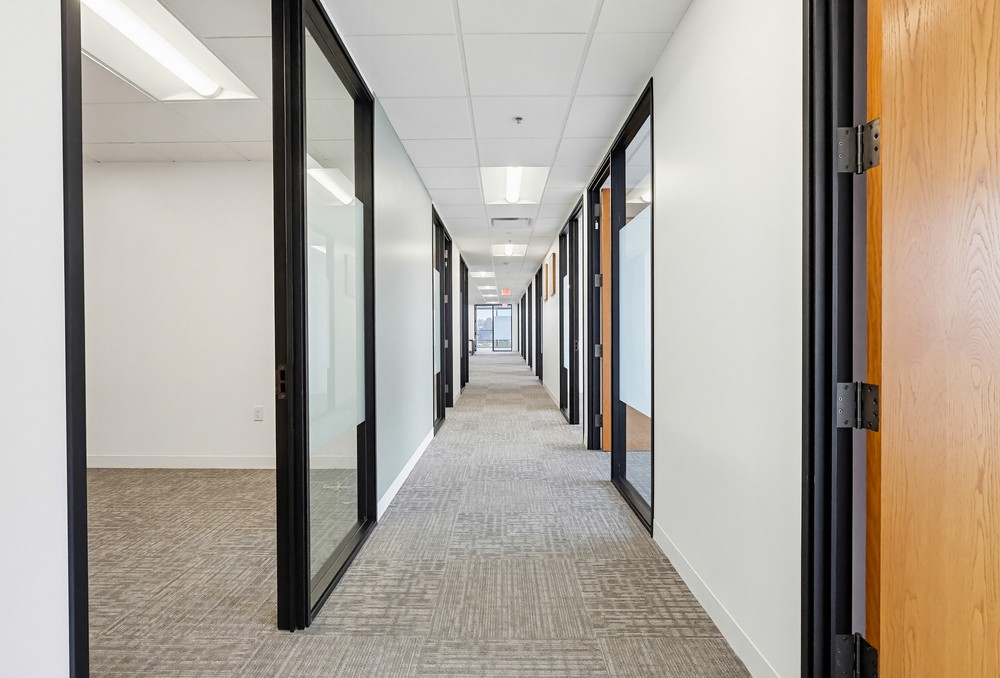 Barrister Suites Office Lease A bright, modern office hallway with beige carpet, white walls, glass office doors, and a ceiling with fluorescent lighting. The hallway extends into the distance, with offices on both sides.