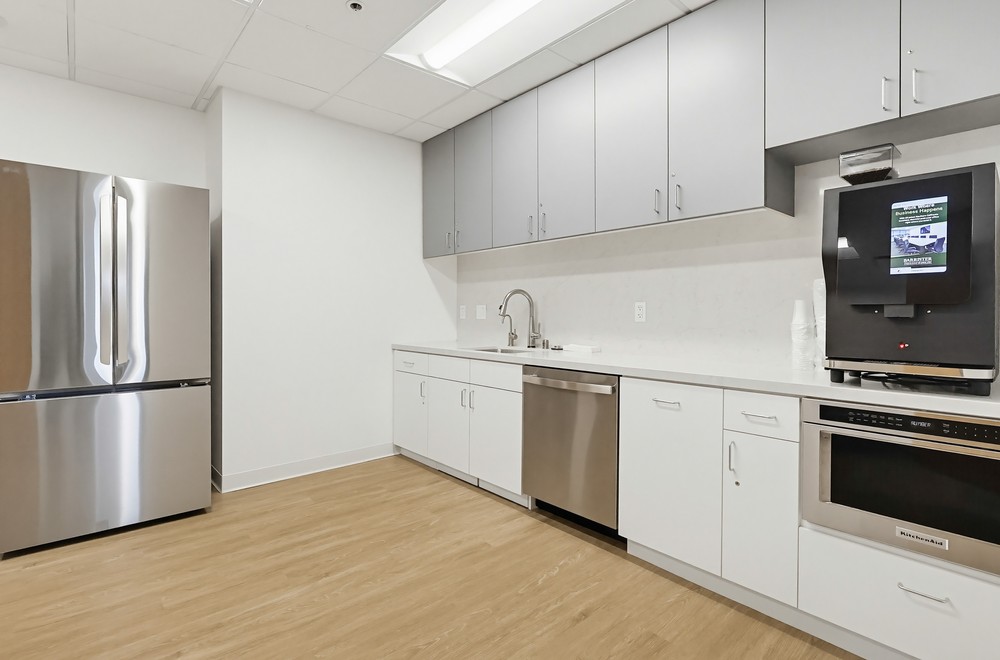 Barrister Suites Office Lease Modern office kitchen with light wood flooring, white cabinets, stainless steel refrigerator and dishwasher, microwave, and a coffee machine on the countertop under wall cabinets.