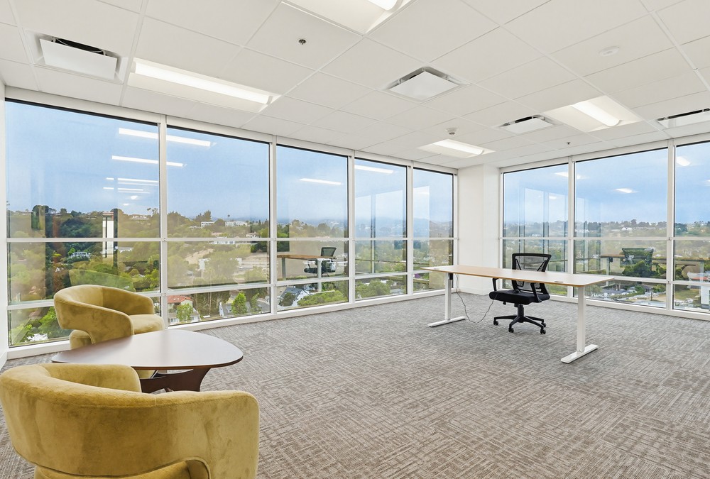Barrister Suites Office Lease Modern office space with large floor-to-ceiling windows, an empty desk with a chair, and two yellow armchairs around a small table. Outside the windows is a scenic view of greenery and hills.