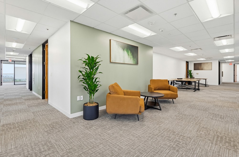 Barrister Suites Office Lease Modern office space with two tan armchairs and a round table on a carpeted floor, a green accent wall with art, potted plant, and long conference tables in the background under bright ceiling lights.