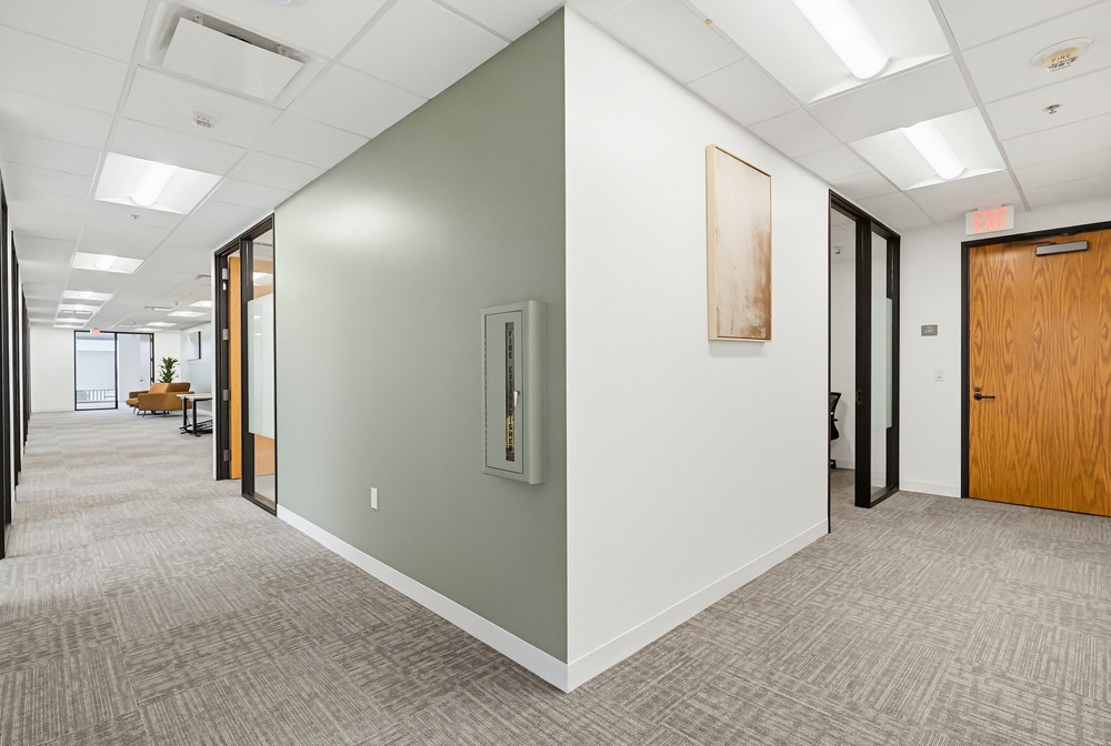 Barrister Suites Office Lease A modern office hallway with neutral colors, beige carpet, white and sage green walls, wooden doors, and glass office partitions. A framed art piece and a utility panel are mounted on the walls. Ceiling lights provide bright illumination.