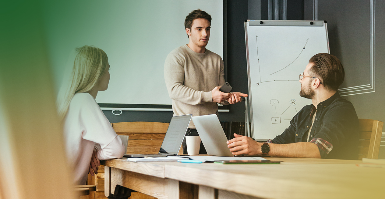 Barrister Suites Office Lease Three people in a meeting room; one man stands by a flip chart with a graph, presenting to two colleagues seated at a table with laptops and coffee cups.