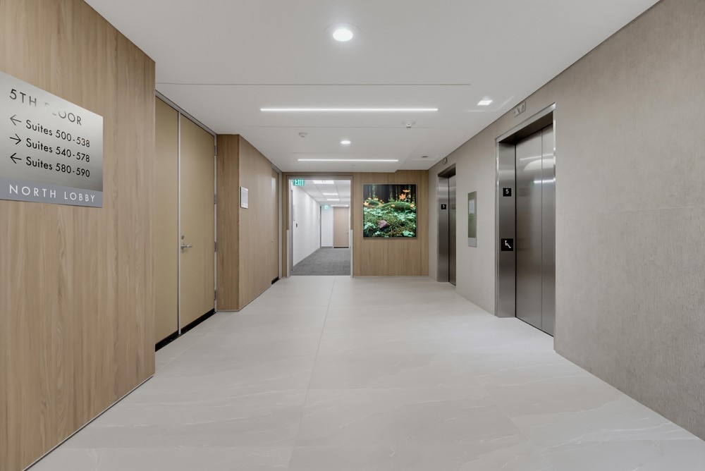 Barrister Suites Office Lease Modern office building hallway with light tile floors, light wood and beige walls, two elevators on the right, and a sign indicating suite numbers on the left. A green exit sign and a wall display with plants are visible ahead.
