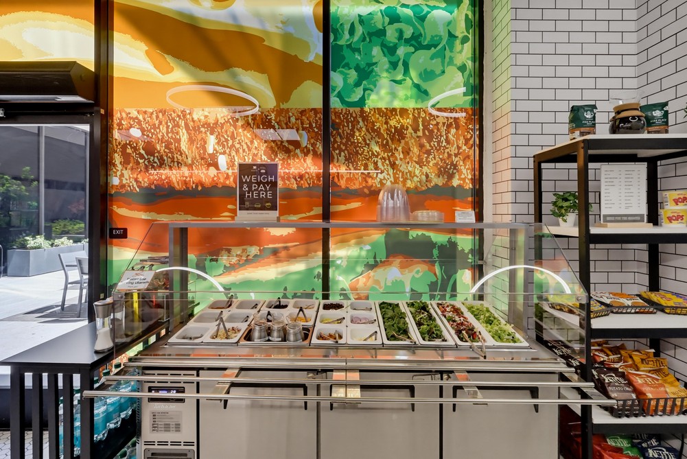 Barrister Suites Office Lease A salad bar with various fresh ingredients is set against a colorful, abstract wall mural. Shelves with snacks appear on the right, and sunlight streams in from windows on the left.