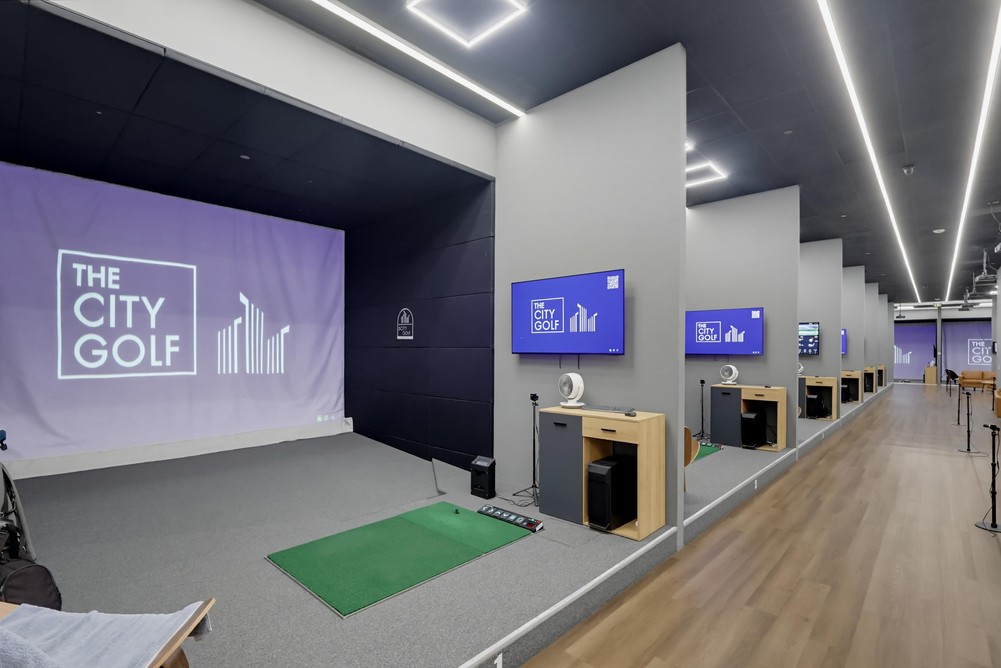 Barrister Suites Office Lease A modern indoor golf simulator facility with several individual stations, each featuring a screen, golf mat, and equipment. The main screen displays The City Golf. The space has sleek, minimalist design and bright lighting.