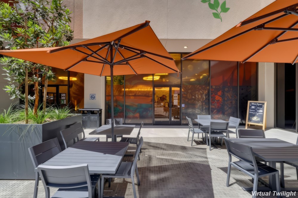 Barrister Suites Office Lease Outdoor patio with gray tables and chairs under large orange umbrellas, next to a building with large windows, greenery, and a chalkboard sign by the entrance.