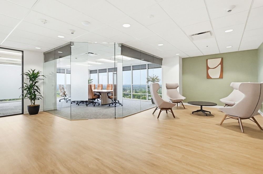 Barrister Suites Office Lease Modern office with light wood flooring, two beige lounge chairs, a round black table, potted plants, and a glass-walled conference room with a large table and orange chairs. Large windows provide natural light and outdoor views.