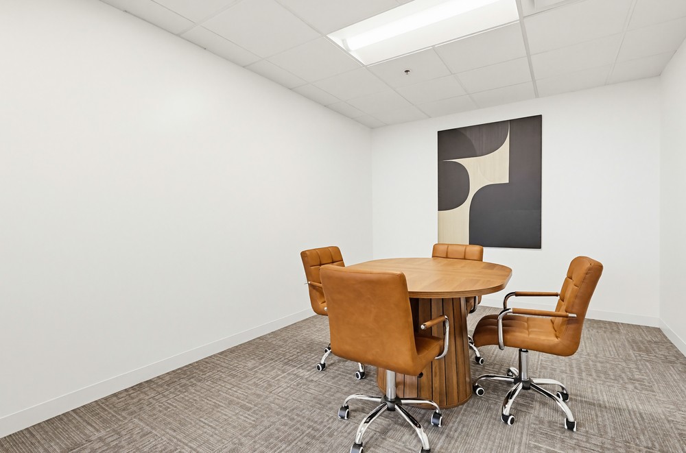 Barrister Suites Office Lease A small conference room with four brown leather chairs around a round wooden table, beige carpet, white walls, and a modern abstract painting on the wall.