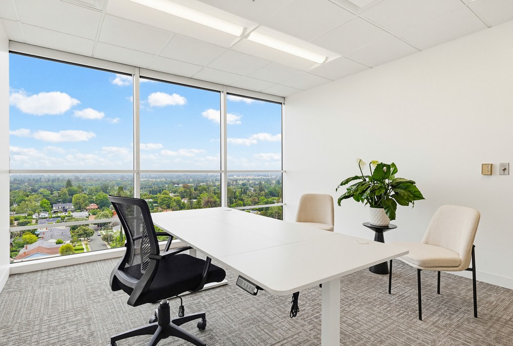 Barrister Suites Office Lease Modern office with a white desk, black office chair, two beige chairs, a potted plant, and large windows offering a scenic view of trees and sky.