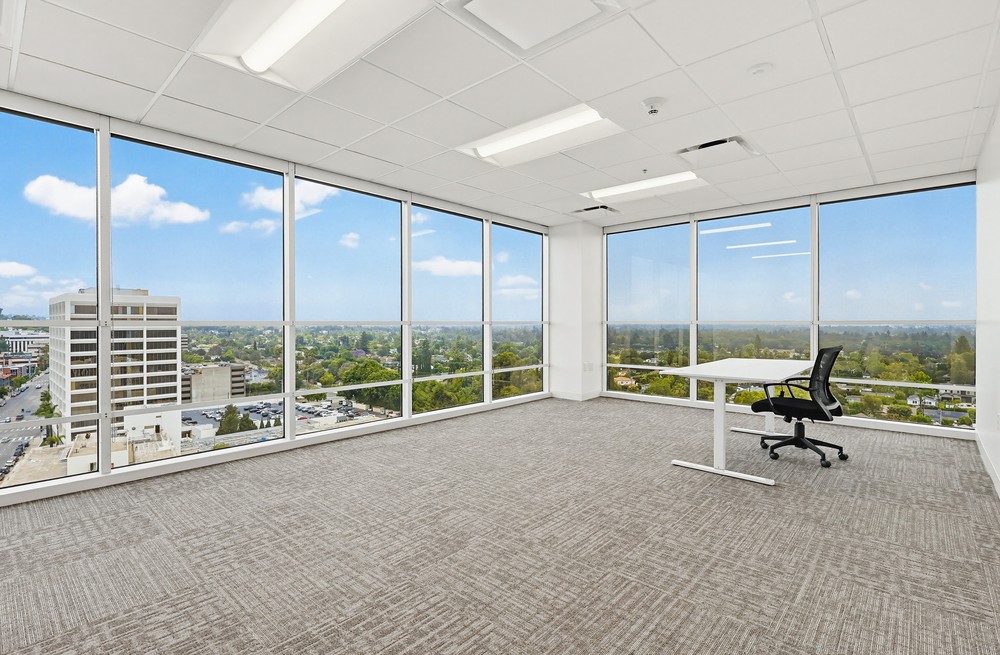 Barrister Suites Office Lease A modern, empty office with large floor-to-ceiling windows, a single desk, and an office chair. The windows offer a cityscape view with buildings, trees, and a blue sky with clouds.