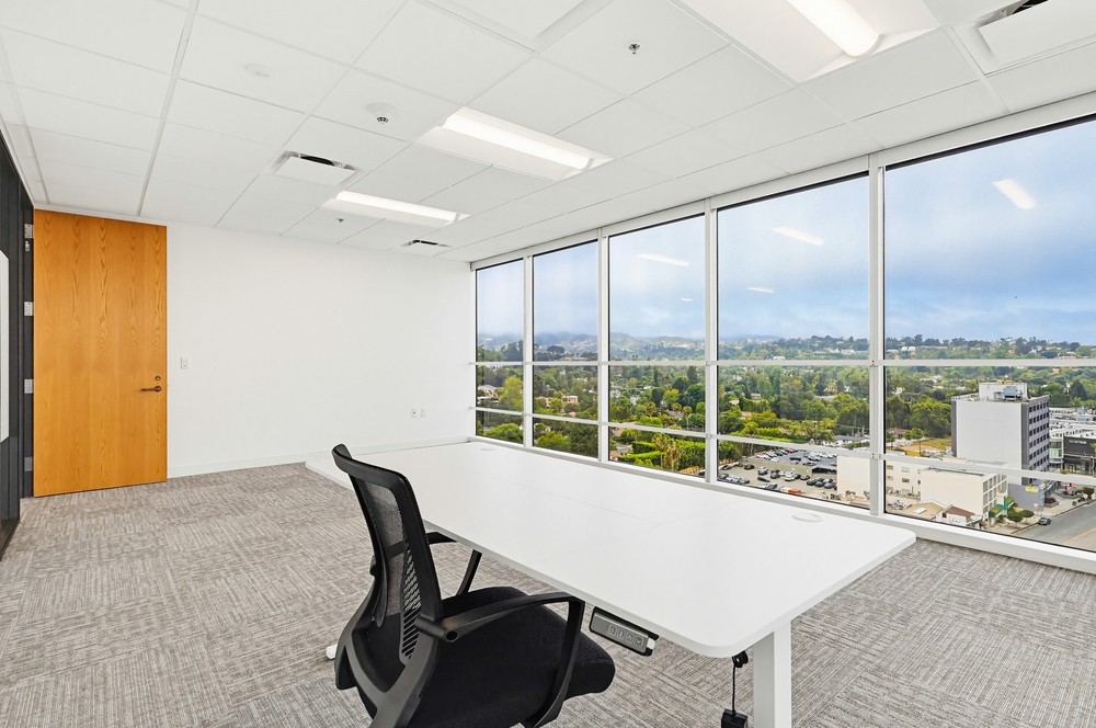 Barrister Suites Office Lease Modern office with a single desk and chair near large windows overlooking a cityscape and greenery, with natural light brightening the clean, minimal space.