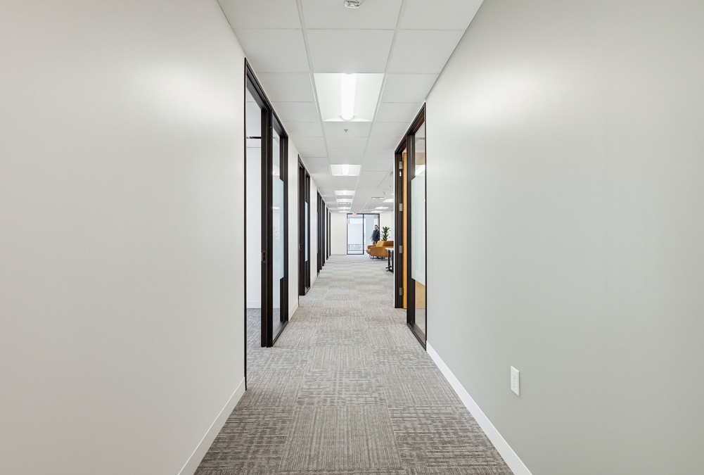 Barrister Suites Office Lease A bright hallway in a modern office building with gray carpet, white walls, and doors leading to individual offices on both sides. The end of the hallway is visible, with a desk and chair in the distance.