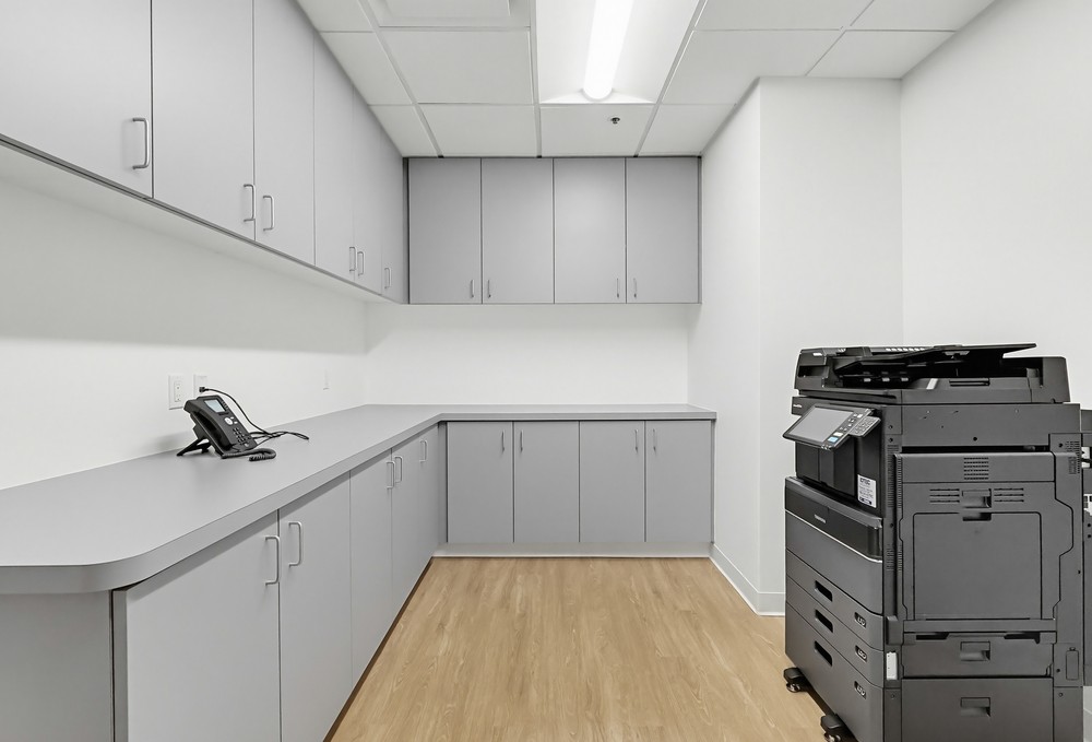 Barrister Suites Office Lease A modern office copy room with gray cabinets, a wood floor, a black copier/printer, and a desk phone, all under bright fluorescent lighting.