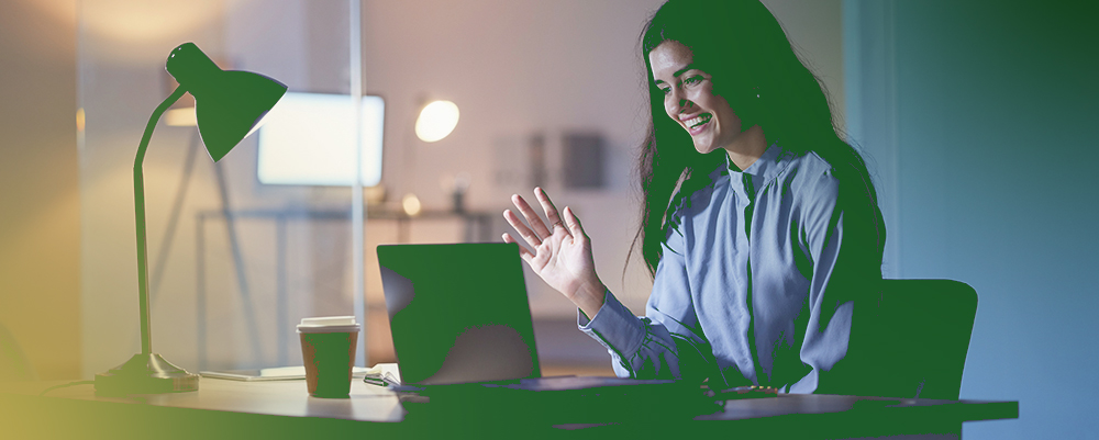 Barrister Suites Office Lease A woman sits at a desk with a laptop, smiling and waving during a video call. A desk lamp, coffee cup, and office background are visible. The setting suggests she is working late.
