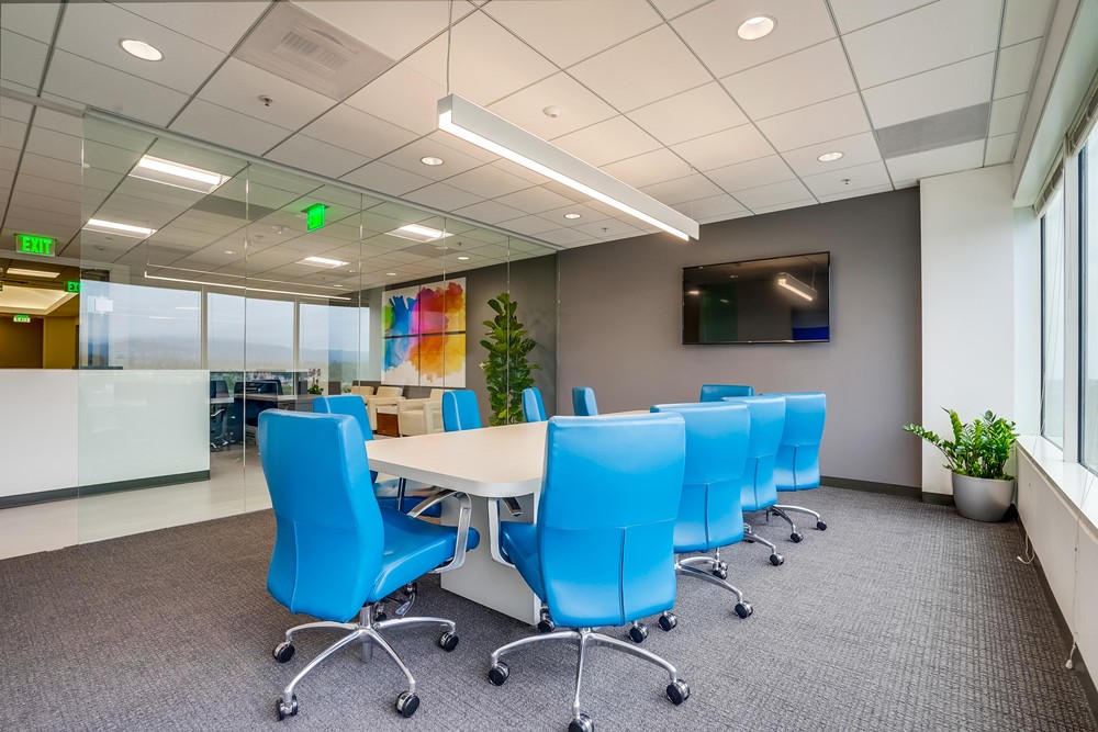 Barrister Suites Office Lease Modern conference room with bright blue chairs around a table, large windows, wall-mounted TV, indoor plants, and glass walls showing an adjacent office area.