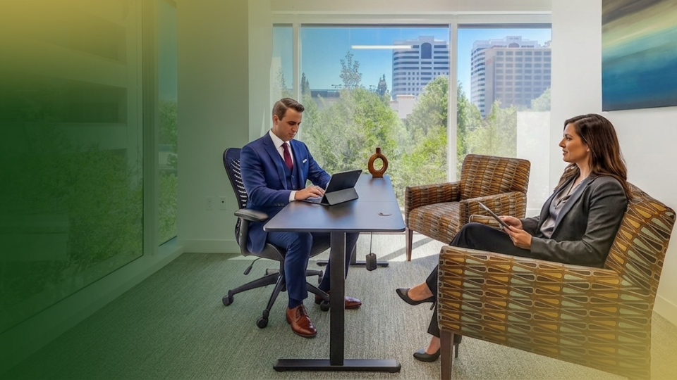 Barrister Suites Office Lease Two people in business attire sit in a modern office with large windows. One types on a laptop at a desk, while the other sits across holding a tablet. City buildings and trees are visible outside.