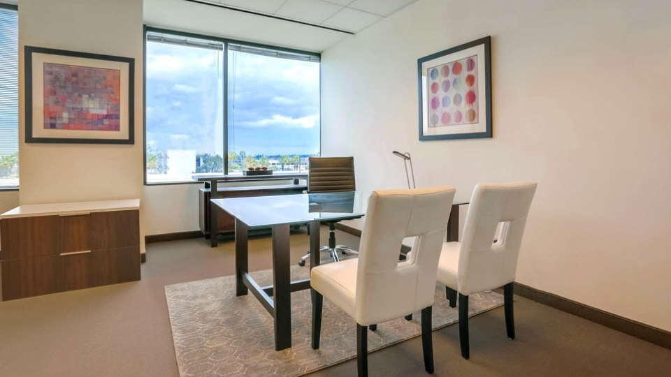 Barrister Suites Office Lease Modern office with a glass desk, leather chair, two white chairs, large window, abstract art on walls, and a cabinet under the window.