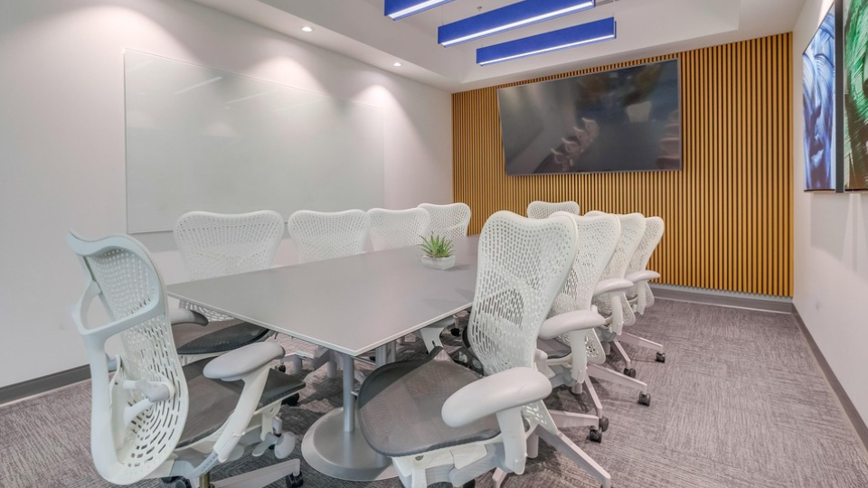 Barrister Suites Office Lease Modern conference room with a long table, ten white mesh chairs, a wall-mounted TV, a whiteboard, wood paneling, and a small plant centerpiece. The room has gray carpet and overhead blue lights.