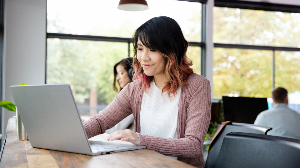 Barrister Suites Office Lease A woman with shoulder-length hair works on a laptop at a wooden table in a bright, modern office with large windows. Other people are working in the background.