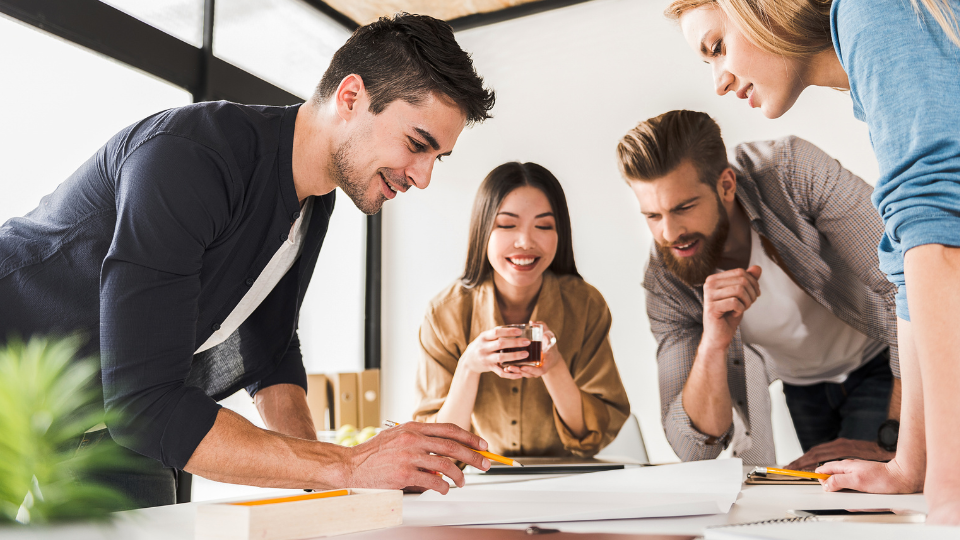 Barrister Suites Office Lease Four young adults gather around a table, smiling and collaborating on a project. One person writes while others watch and discuss, creating a positive, engaging atmosphere in a modern office space.