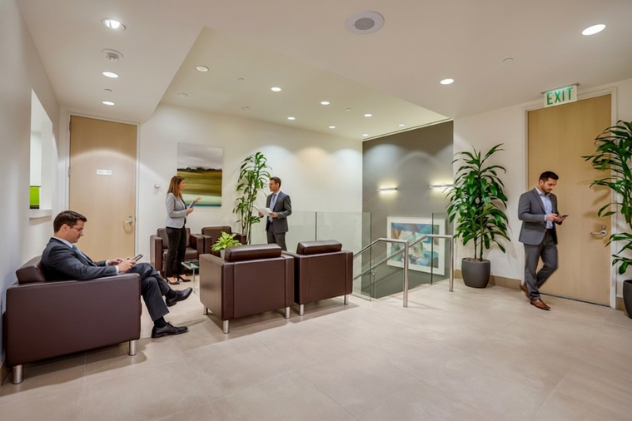 Barrister Suites Office Lease Four people in business attire are in a modern office lobby with brown chairs, large green plants, and neutral decor. Two are seated on their phones; two are standing and talking near a painting and staircase.