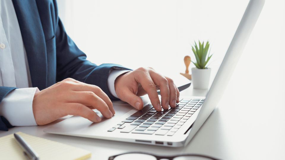 Barrister Suites Office Lease A person in a suit types on a laptop at a white desk with a notepad, pen, glasses, and a small potted plant nearby.