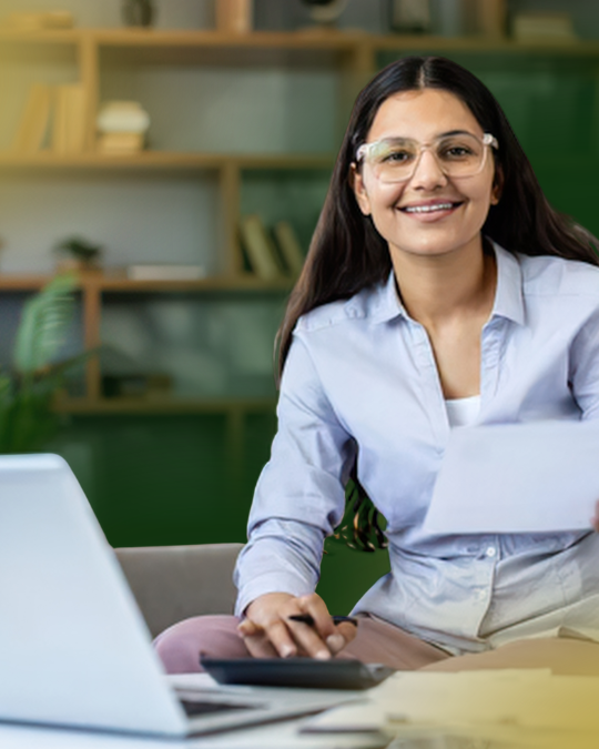 Barrister Suites Office Lease A woman with long brown hair and glasses sits at a desk with a laptop, holding a piece of paper and smiling, with shelves and plants in the background.