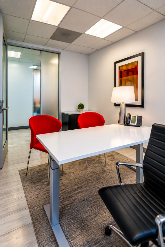 Barrister Suites Office Lease Modern office with a white desk, black office chair, two red guest chairs, a lamp, phone, framed art, and small plant on a cabinet. Glass door and wall in the background.