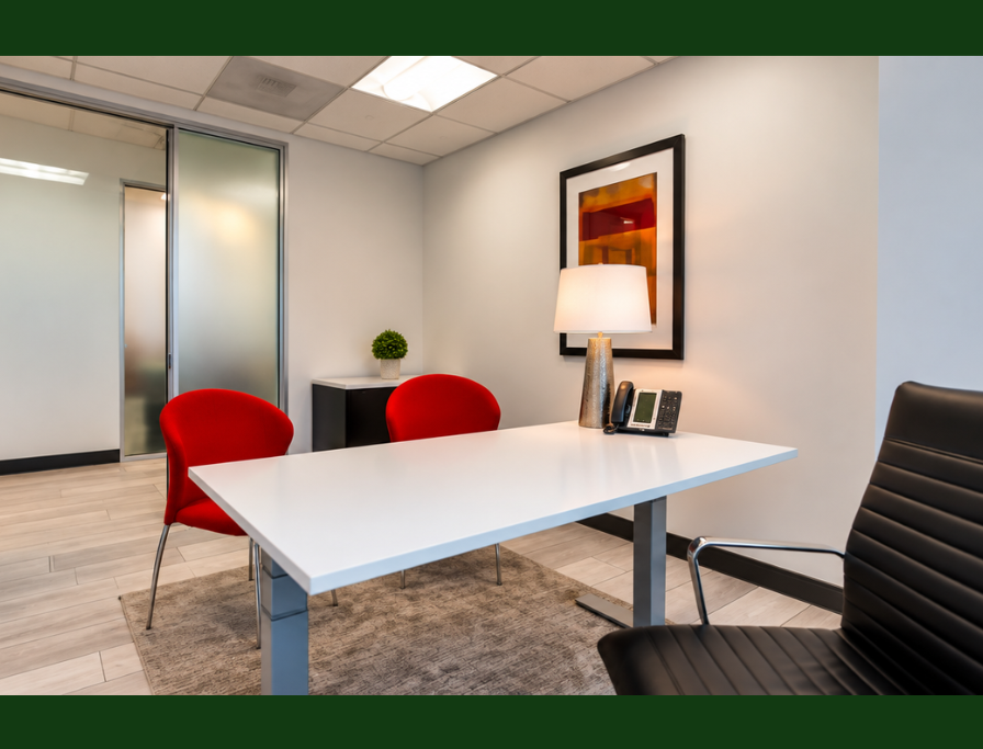 Barrister Suites Office Lease Modern office with a white desk, black chair, two red chairs, a table lamp, phone, artwork on the wall, and a small potted plant by a glass door. Neutral walls and wood floor create a clean, professional look.