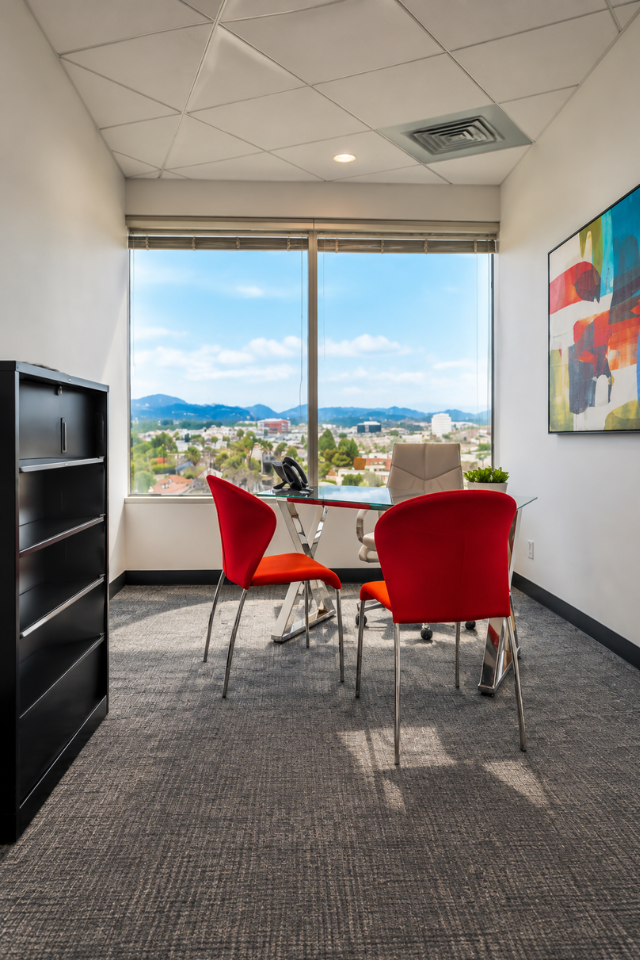 Barrister Suites Office Lease Modern office with two red chairs and a glass desk facing a large window with a scenic city and mountain view. Abstract art hangs on the wall, and a black shelf stands on the left. Bright, clean, and professional atmosphere.