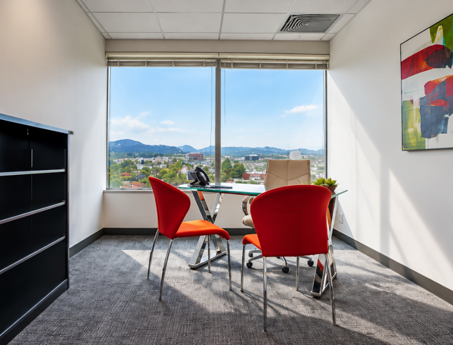 Barrister Suites Office Lease Modern office with a glass desk, tan chair, two red chairs, phone, plant, abstract wall art, and large window showing a cityscape and mountains under a blue sky.
