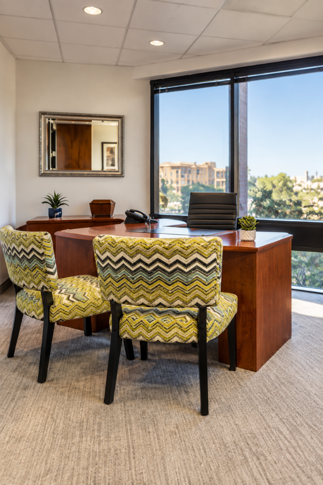 Barrister Suites Office Lease Modern office with large windows, a wooden desk, black office chair, two patterned guest chairs, a mirror, and small decor items on the desk. Cityscape visible outside.