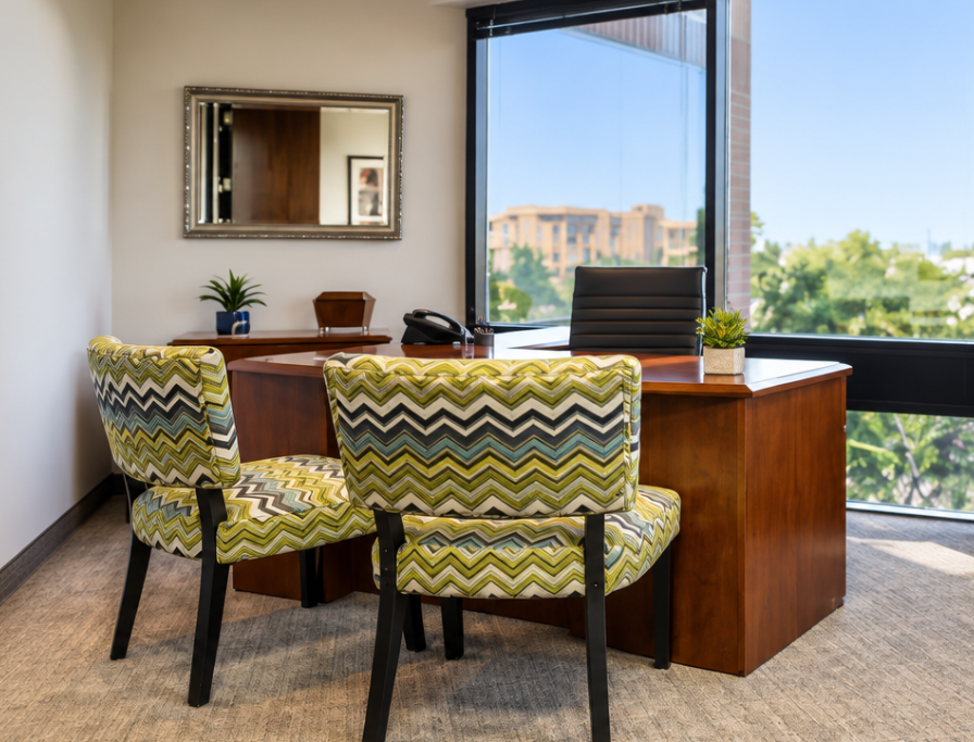 Barrister Suites Office Lease A modern office features a wooden desk, a black chair, and two patterned guest chairs. A window provides a view of buildings and trees. A small plant and a mirror decorate the space.