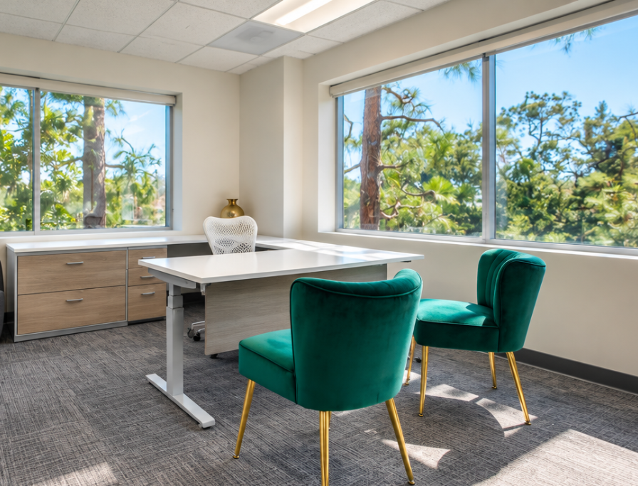 Barrister Suites Office Lease Modern office with a white desk, two green velvet chairs, a white office chair, and large windows showcasing trees and greenery outside. Light gray carpet and neutral walls complete the bright, airy space.