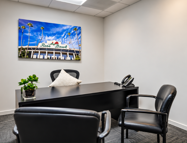 Barrister Suites Office Lease Modern office with a black desk, two black chairs, a potted plant, a pillow, a desk phone, and a framed photo of the Rose Bowl stadium on the wall. The room has white walls and a gray carpet.