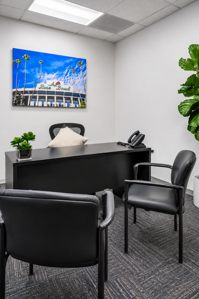 Barrister Suites Office Lease A modern office with a black desk, two black chairs, a potted plant, phone, and a framed photo of the Rose Bowl stadium on the wall. The room has gray carpet and white walls.