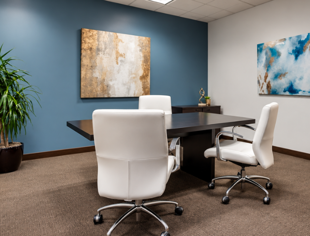 Barrister Suites Office Lease Modern office with a dark wood desk, two white chairs, blue accent wall, abstract art, and a potted plant in the corner. The space is clean and well-lit.