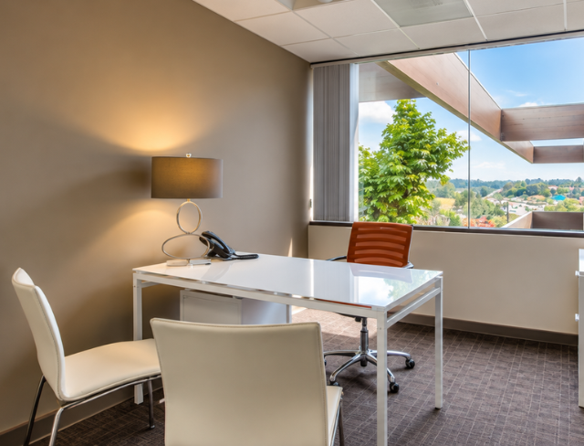 Barrister Suites Office Lease Modern office with a white desk, two chairs, a lamp, and a phone. Large window with a view of trees and buildings outside. Neutral walls and carpeted floor.