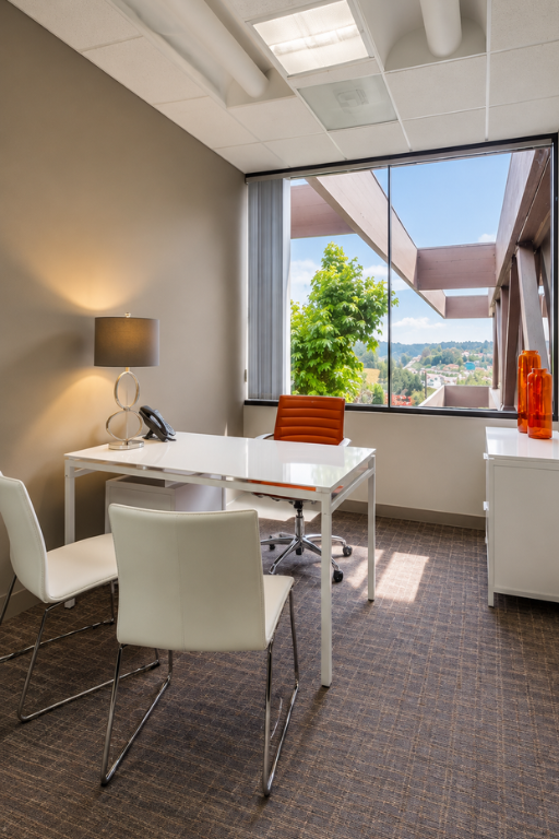 Barrister Suites Office Lease Modern office with a white desk, orange chair, two white guest chairs, lamp, phone, and a large window showing trees and a view outside. Two orange vases sit on a white cabinet by the window.