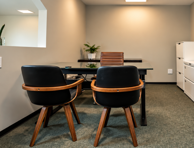 Barrister Suites Office Lease Modern office with a glass desk, a brown leather chair behind it, and two black chairs with wooden legs in front. A plant sits on the desk, and white cabinets are against the wall.