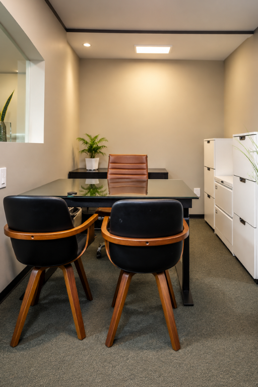 Barrister Suites Office Lease A modern office with a glass desk, brown leather chair behind it, two black chairs in front, white filing cabinets, and green plants. The space is well-lit and has a minimalist design.