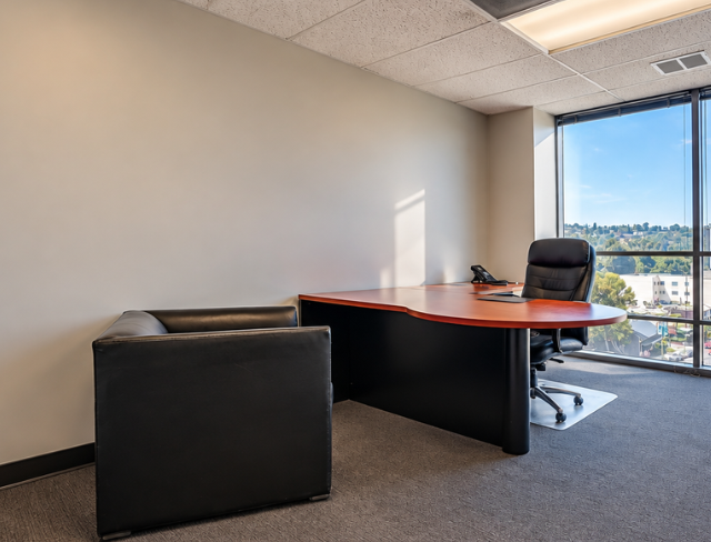 Barrister Suites Office Lease Modern office with a large L-shaped desk, black chair, leather guest chair, telephone, and bright window overlooking a cityscape. Walls are beige and the floor is carpeted.