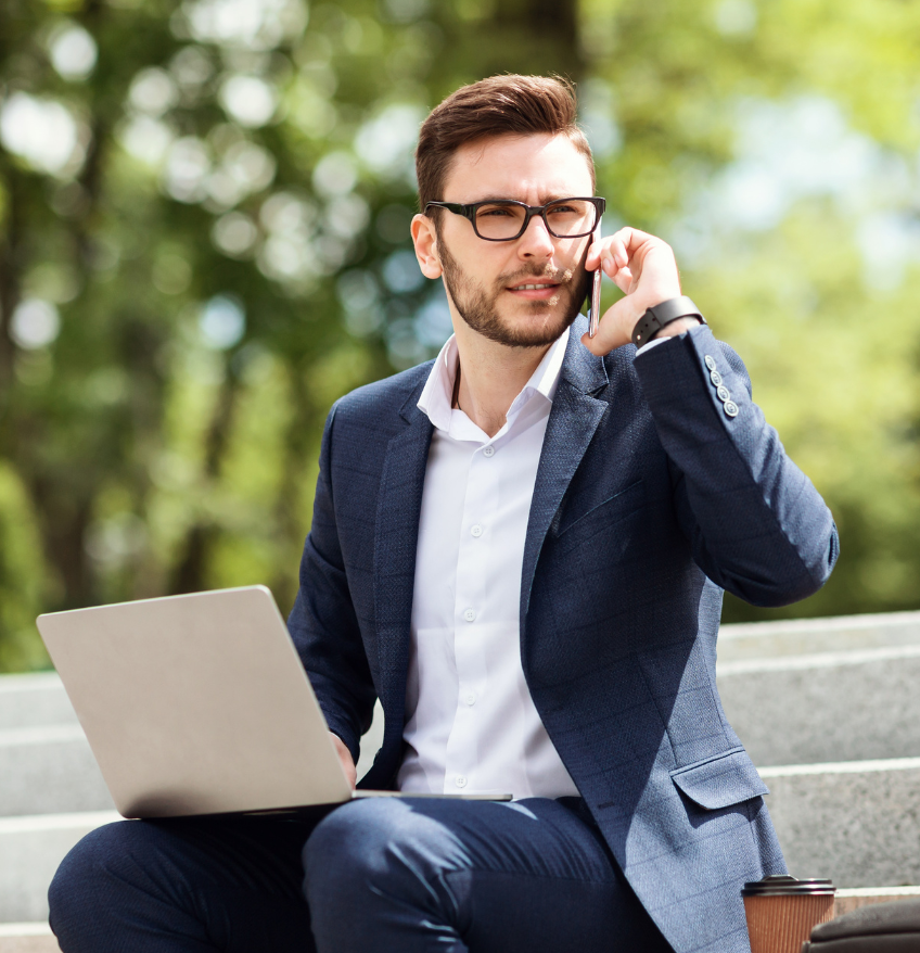 Barrister Suites Office Lease A man in a suit sits on outdoor steps, holding a laptop on his lap and talking on a smartphone. He wears glasses and looks focused. Green trees are blurred in the background.