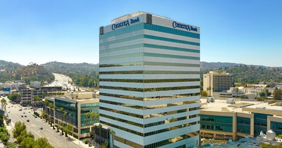 Barrister Suites Office Lease A tall office building with Comerica Bank signage stands among other buildings and roads, with trees and hills in the background under a clear sky.