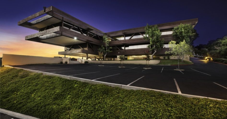 Barrister Suites Office Lease A modern, angular office building with large overhangs and glass windows is shown at dusk, with lights on inside. The parking lot in front is empty, and there are trees and green landscaping in the foreground.